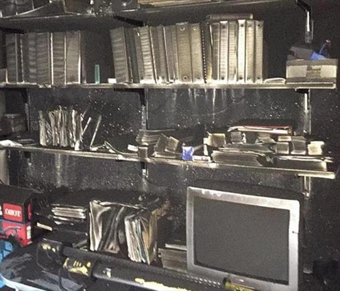 A computer on a desk top, books on shelves, and the wall behind them all covered in heavy soot.